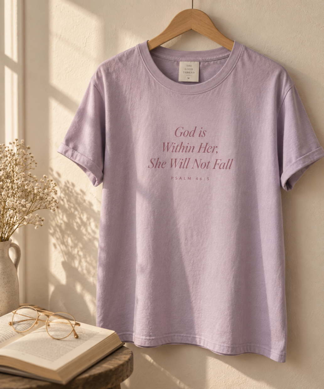 Soft Lavender relaxed-fit tee printed with “God is Within Her, She Will Not Fall” in mauve serif type, folded on a linen backdrop